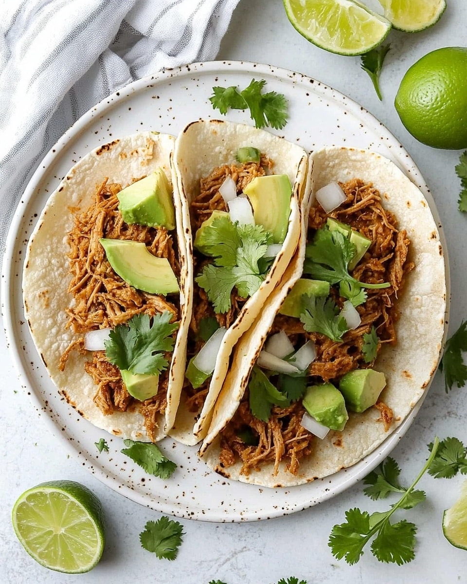 Shredded Chicken Street Tacos, Mexican street taco recipe, easy shredded chicken tacos, quick taco dinner, flavorful chicken tacos Four small soft white corn tortillas are placed side by side on a round white plate. Each tortilla holds a layer of shredded cooked chicken with a light brown color, topped with small cubes of green avocado and bits of white onion. On top of the avocado and onion, there are a few fresh green cilantro leaves scattered on each taco. Around the plate, there are two bright green lime wedges, one whole green lime, and sprigs of cilantro for garnish. The plate is set on a white marbled surface with a white cloth napkin with gray stripes partially visible on the bottom right.