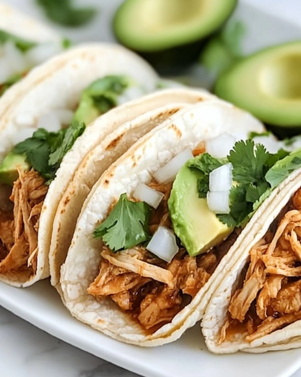 Shredded Chicken Street Tacos, Mexican street taco recipe, easy shredded chicken tacos, quick taco dinner, flavorful chicken tacos The image shows three soft tacos placed side by side on a white plate. Each taco has a light golden tortilla wrapped around a filling of shredded brownish cooked meat. On top of the meat in each taco is a layer of small bright green avocado cubes and chopped white onion pieces. Fresh green cilantro leaves are spread across the tacos, adding a pop of color. In the background, there are two lime wedges, one slightly blurred, resting on a white marbled surface. The overall look is fresh and vibrant, with the soft textures of the tortillas and meat contrasting the crispness of the avocado and onions.