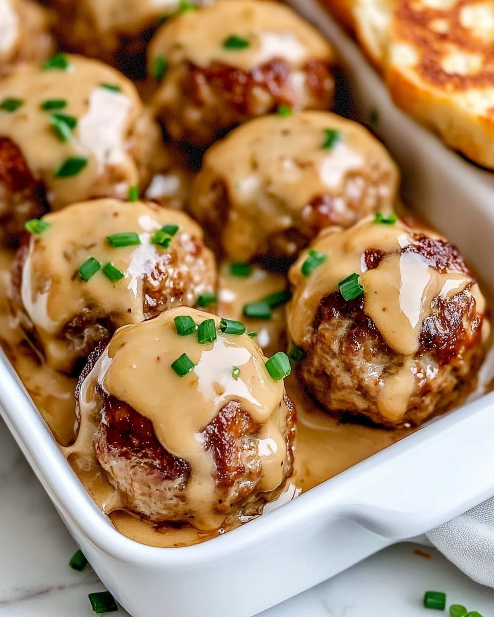 Rotel Cream Cheese Sausage Balls, cheesy sausage bites, easy party appetizers, flavorful sausage snacks, crowd-pleasing sausage balls The dish shows a white rectangular dish filled with two layers of browned meatballs that have a slightly rough texture. Each meatball is covered with thick light yellow sauce that looks smooth and creamy, dripping slightly down the sides. The top layer of meatballs is sprinkled with small pieces of fresh green chives, adding a fresh touch. The dish sits on a white marbled surface, and a slice of light brown toasted bread is partially visible on the right side.