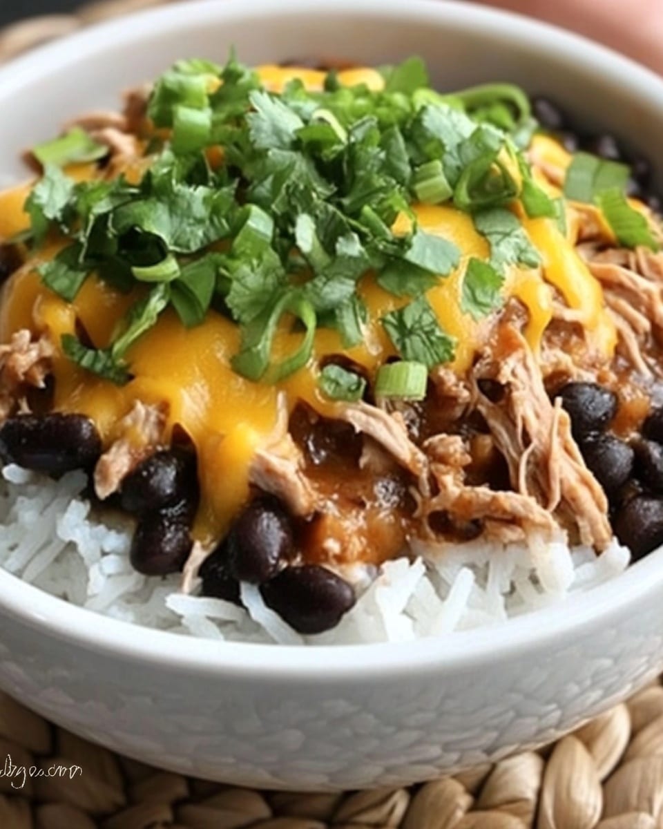 Crock Pot Santa Fe Chicken, slow cooker Santa Fe chicken, easy Santa Fe chicken recipe, flavorful chicken casserole, healthy slow cooker dinner The image shows a black bowl filled with white rice at the bottom. On top of the rice is a generous layer of shredded cooked meat mixed with black beans. The meat layer is topped with melted yellow cheese, and the dish is garnished with chopped green onions scattered across the top. The black bowl sits on a woven brown mat, set against a white marbled surface.