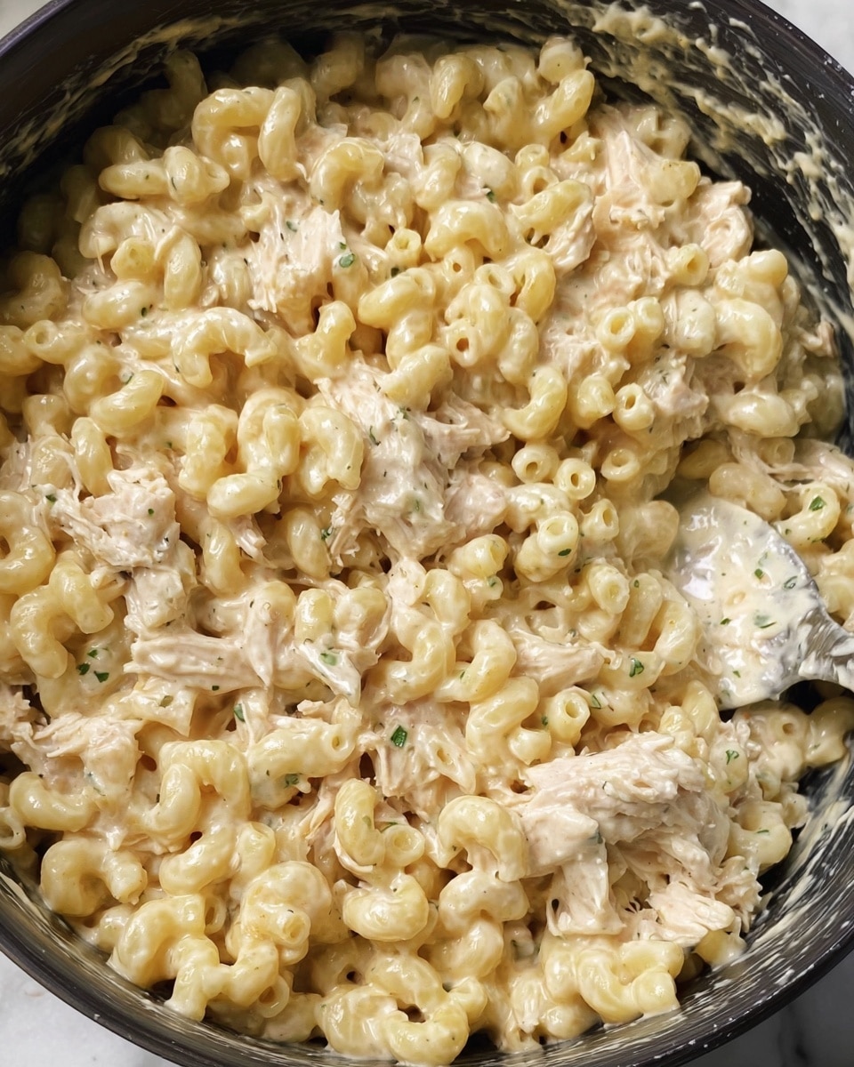 A close-up view of a creamy macaroni dish in a dark pot against a white marbled background, showing a single layer of pale yellow elbow macaroni mixed thickly with shredded, light beige chicken and a smooth, glossy white sauce with small green herb pieces. The texture looks soft and rich, with sauce coating each pasta and chicken part evenly, giving a dense and comforting appearance. A metal spoon covered partly in sauce is on the side, blending gently into the food. photo taken with an iphone --ar 4:5 --v 7