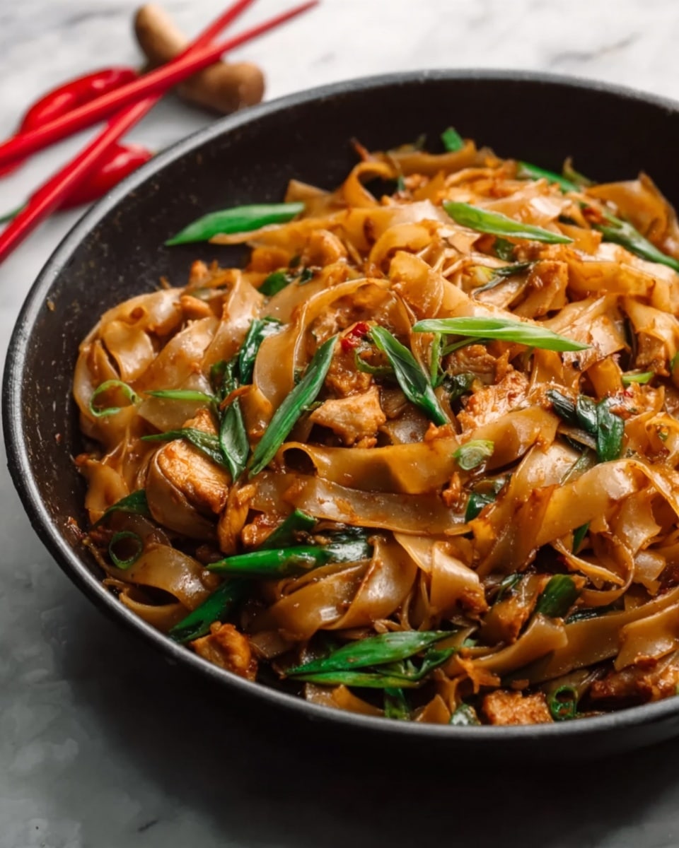 A close-up view of a black pan filled with wide, flat noodles that are glossy brown, mixed with pieces of golden-brown chicken and bright green chopped scallions scattered on top. The noodles look saucy and shiny, showing a rich, savory coating. The pan sits on a white marbled surface with a pair of red chopsticks beside it. photo taken with an iphone --ar 4:5 --v 7