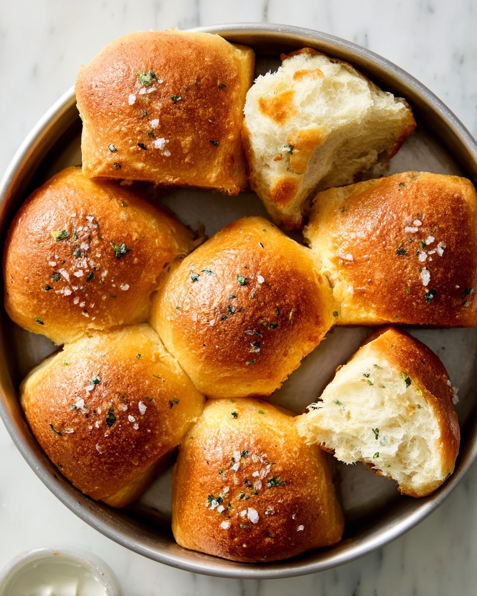 A round baking pan holds seven golden brown dinner rolls arranged in a circle with one roll slightly pulled away, showing soft, fluffy white bread inside. The rolls have a shiny, slightly crispy top sprinkled with coarse sea salt and small green herb bits. The surface of each roll is smooth with a rich, warm brown color and a light texture that suggests softness underneath. The pan rests on a white marbled surface. Photo taken with an iphone --ar 4:5 --v 7