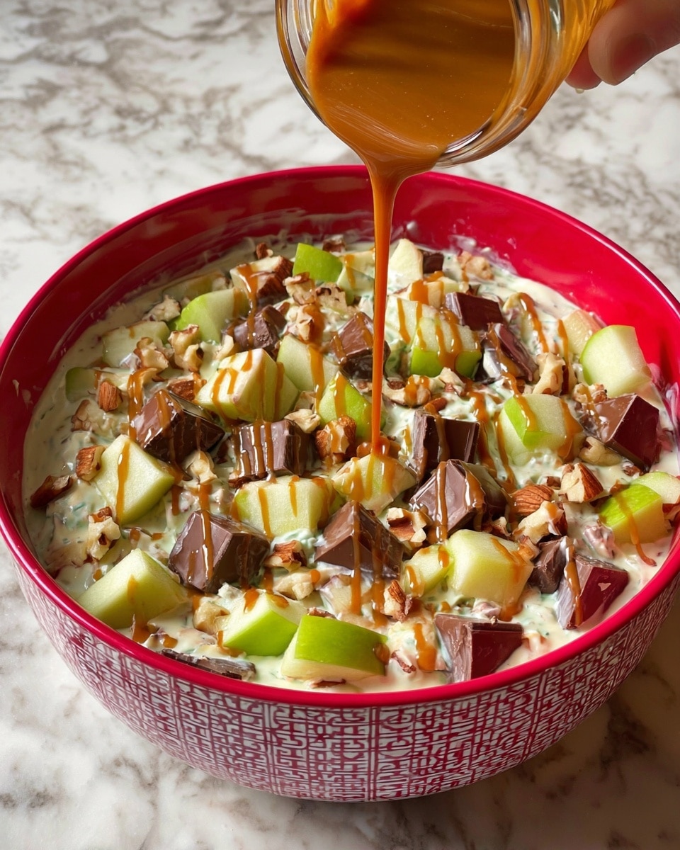 A red bowl with white chain pattern holds a creamy base mixed with chunks of green apple and chopped chocolate candy pieces with nuts. The creamy layer is off-white and thick, filling the bowl almost to the top. On top, there are larger pieces of bright green apple and chunky chocolate candy pieces scattered evenly around. A caramel-colored sauce is being poured from a jar held by a woman's hand onto the top, creating thin, shiny drizzles over the whole dish. The bowl is on a white marbled surface. photo taken with an iphone --ar 4:5 --v 7