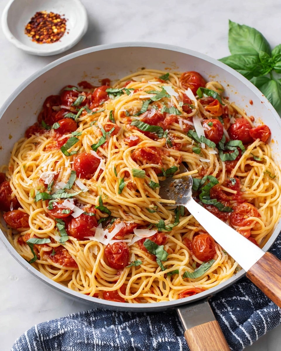 A close-up of a white pan filled with spaghetti mixed with a red tomato sauce, showing uneven small chunks of cooked tomatoes and fresh green basil leaves scattered on top. Thin spaghetti strands have a glossy texture from the sauce, and some thin pale yellow cheese shavings are sprinkled over the dish. A woman's hand is holding a fork twirling some spaghetti with sauce and tomato pieces lifted above the pan. The pan sits on a white marbled surface beside a white bowl. photo taken with an iphone --ar 4:5 --v 7