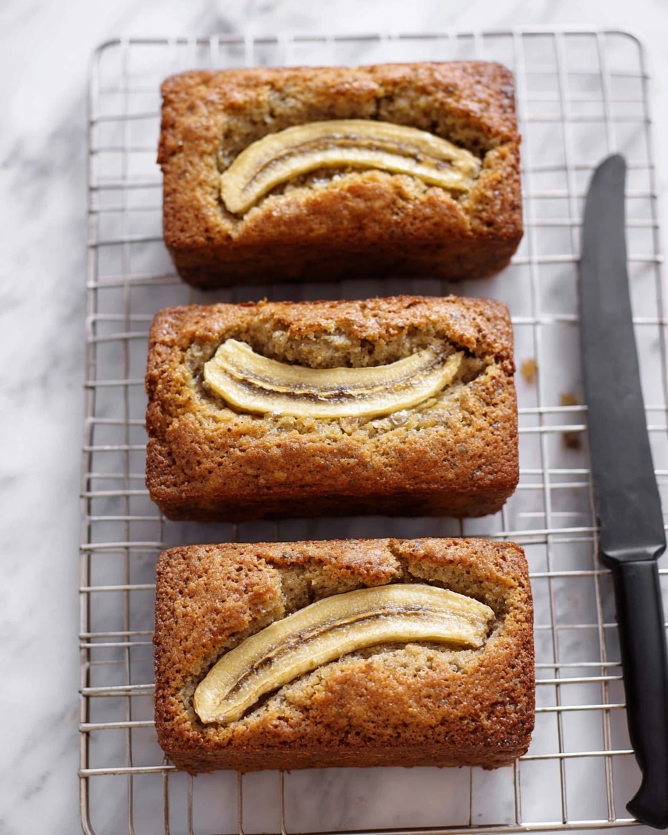 Three small rectangular banana breads sit on a silver wire cooling rack over a white marbled surface. Each bread is a golden brown color with a rough, slightly bumpy texture and has a single half-slice of banana laid across the top, showing a creamy yellow and light brown color. To the right of the breads, there is a long black-handled knife placed on the white marbled surface. Photo taken with an iphone --ar 4:5 --v 7