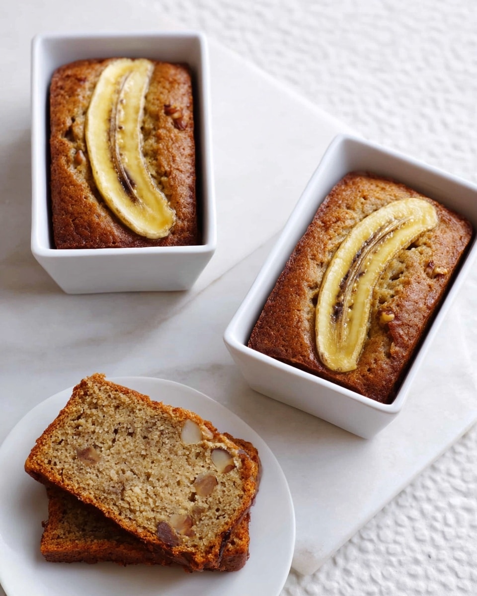 Two small rectangular banana breads with a golden-brown crust are in white square ramekins, each topped with a split banana slice showing light yellow and brown colors. Nearby, a white round plate holds three slices of banana bread stacked together, showing a soft light brown interior with walnut bits. One extra slice of banana bread lies flat on the white marbled surface, revealing its texture and scattered nuts inside. photo taken with an iphone --ar 4:5 --v 7
