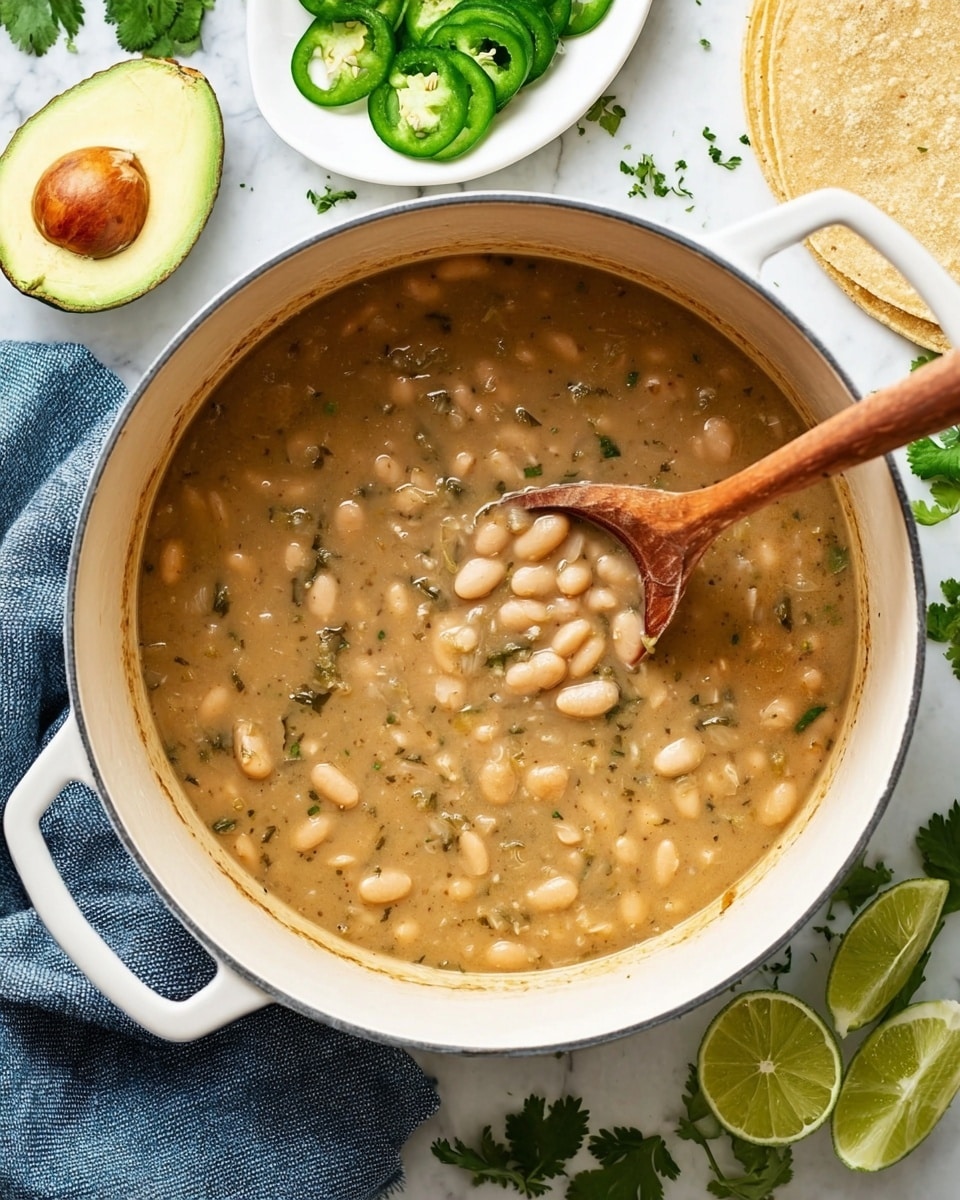 The image shows a white pot filled with creamy, light brown bean soup that has visible white beans and small green vegetable bits mixed in. A wooden spoon is scooping some of the soup from the pot. Around the pot, there are lime slices at the bottom right, fresh green cilantro leaves, a blue cloth napkin on the left, half an avocado with its seed on the top left, sliced green jalapeño peppers on a white plate at the top center, and a beige tortilla on the upper right. All items are placed on a white marbled surface. photo taken with an iphone --ar 4:5 --v 7