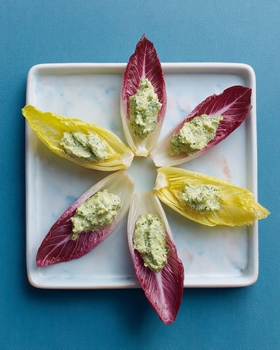 A white square plate holds six endive leaves arranged in a circle, alternating between three yellow-tipped leaves and three dark pink-tipped leaves. Each leaf is filled with a dollop of light green creamy spread with a slightly chunky texture. The plate sits on a white marbled surface, creating a clean and simple background. Photo taken with an iphone --ar 4:5 --v 7