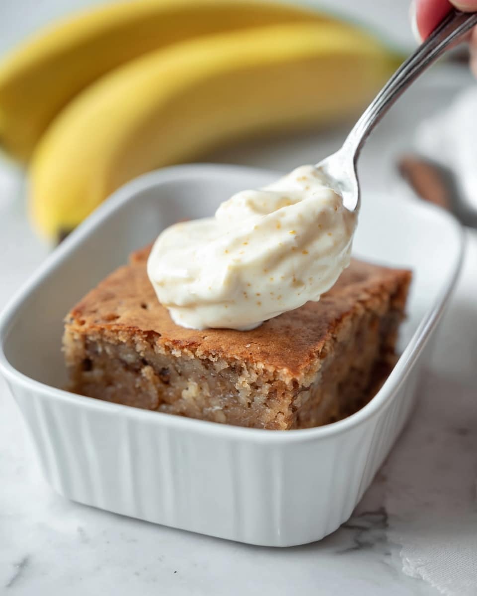 The image shows a white bowl with two layers inside: the bottom layer is a light brown baked square piece with a slightly rough texture, and the top layer is a creamy off-white whipped dollop with soft peaks and tiny specks, placed on the front half of the baked piece. Behind the bowl, there is a white baking dish with more of the light brown baked square and a metal spoon resting inside. Two ripe yellow bananas with small spots lean against the baking dish. The scene is set on a white marbled surface with a shiny metal spoon in the foreground. Photo taken with an iphone --ar 4:5 --v 7