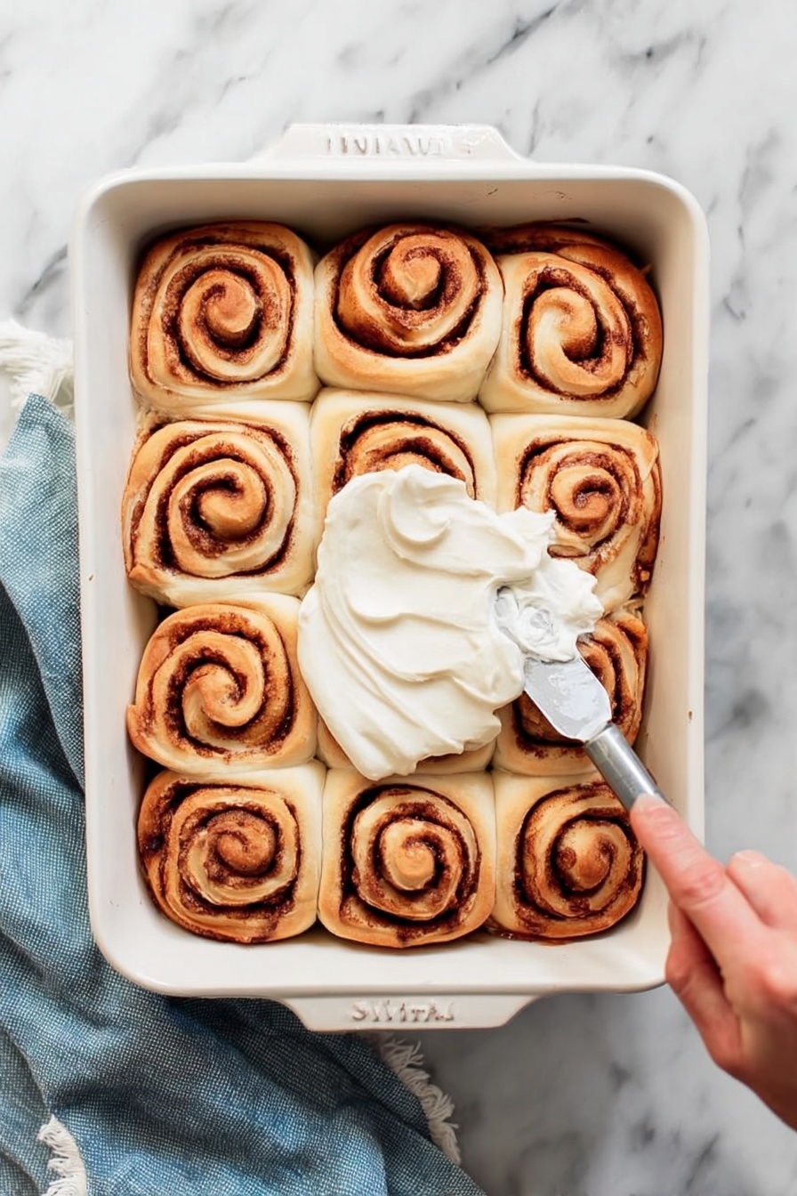 A white rectangular baking dish holds twelve cinnamon rolls arranged in a 3 by 4 grid. Each roll shows a spiral pattern with light brown dough and darker brown cinnamon filling. A woman's hand is spreading smooth white frosting over the center rolls with a silver spreading knife. The dish sits on a white marbled surface with a folded blue cloth nearby. Photo taken with an iphone --ar 2:3 --v 7 - Cinnamon Rolls, Easy Cinnamon Rolls, Soft Cinnamon Rolls, Homemade Cinnamon Rolls, Best Cinnamon Roll Recipe