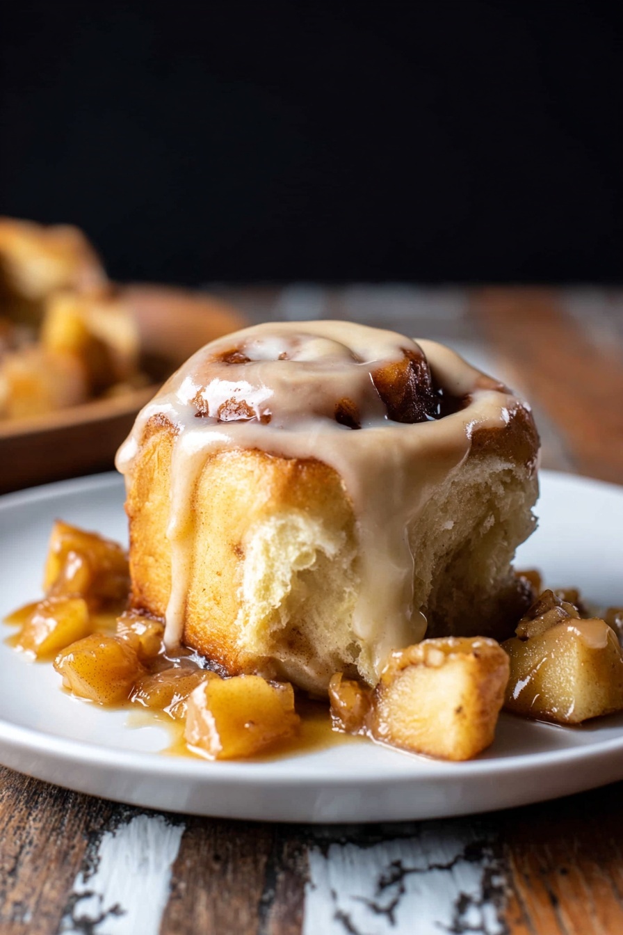 A cinnamon roll with visible swirls and a light golden brown color, topped with thick creamy icing that drapes over the edges, sitting on a white plate. Surrounding the roll are several torn pieces showing a soft, fluffy inside with a slightly darker cinnamon layer. Small chunks of a caramel-like sauce with bits of apple sit around the base, adding a shiny texture. The background is dark, and the plate rests on a rustic wooden surface with a white marbled texture. photo taken with an iphone --ar 2:3 --v 7 - Apple Cinnamon Rolls with Brown Butter Maple Icing, cinnamon apple rolls, fall breakfast recipes, easy cinnamon roll with apple, brown butter maple icing