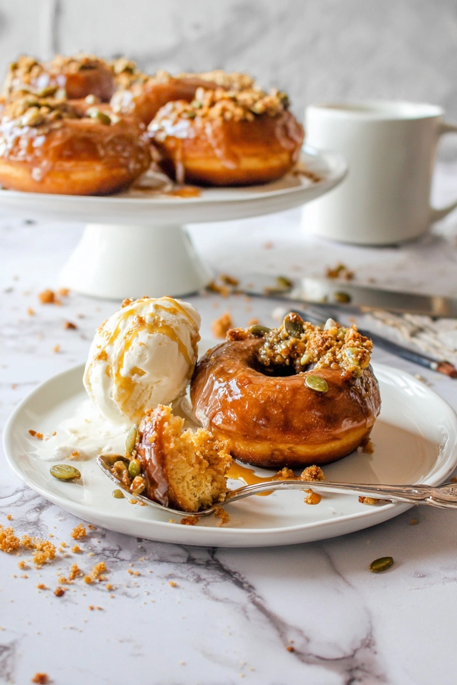 The image shows one brown donut with a shiny glaze, topped with some pumpkin seeds and crumb bits, placed on the right side of a white plate; to its left is a scoop of white ice cream melting slightly with amber syrup drizzled over it, and a spoon in front holds a bite of the donut covered in a mix of ice cream and syrup; in the background there is a white round cake stand holding more donuts with the same toppings, all set on a white marbled surface with scattered syrup drops and crumbs, a silver spoon, and a white mug visible; photo taken with an iphone --ar 2:3 --v 7 - Pumpkin Donuts with Bourbon Caramel Glaze, fall pumpkin donuts, bourbon caramel glaze recipe, homemade pumpkin donuts, autumn dessert ideas
