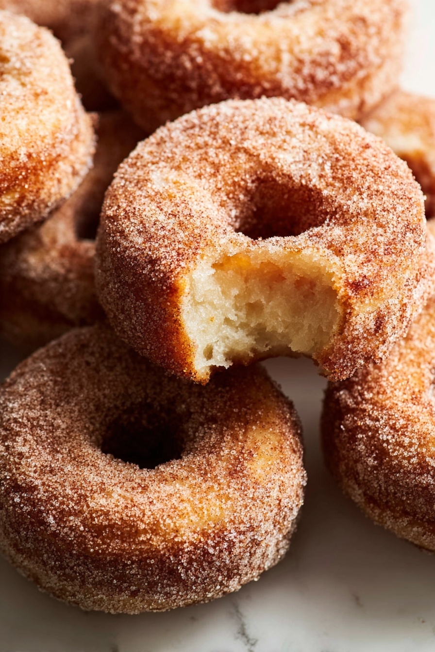 A stack of three sugar-covered donuts sits on a white marbled surface, with the top donut showing a bite taken out, revealing a soft, light brown inside. Each donut has a rough texture coated evenly in sugar, giving a slightly grainy look. The donuts are arranged one on top of the other, with the bitten one on the top, the middle and bottom donuts fully intact, all round with a visible center hole. The lighting softly highlights the sugar crystals and the golden brown color of the fried dough. photo taken with an iphone --ar 2:3 --v 7 - Apple Cider Donuts, homemade apple cider donuts, fall apple donuts, cinnamon sugar donuts, easy apple cider donut recipe