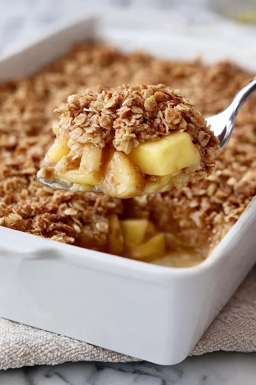 A white rectangular baking dish filled with a dessert that has three visible layers: at the bottom, soft light yellow chunks of apple, in the middle, a slightly sticky cinnamon-spiced syrup, and on top, a thick layer of golden brown oat crumble with a rough texture. A silver spoon is lifting a portion from the dish, showing the layers clearly. The dish is placed on a white marbled surface with a bit of a folded cloth underneath. Photo taken with an iphone --ar 2:3 --v 7 - Amish-Style Apple and Cinnamon Baked Oatmeal, healthy baked oatmeal with apples and cinnamon, cozy breakfast recipes, easy apple breakfast ideas, hearty oatmeal bake