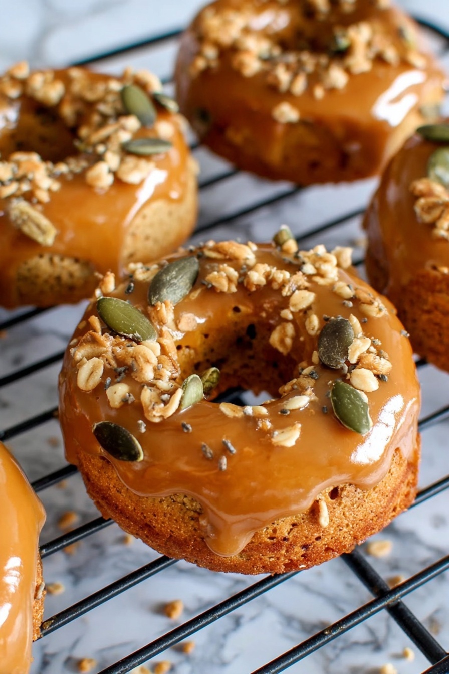 The image shows several small round donuts with a shiny caramel glaze on the top layer, sitting on a black cooling rack. The caramel glaze is smooth and glossy, covering the top and sides of the donuts. On top of the caramel layer, there are scattered crunchy toppings made of light brown crumbles and green pumpkin seeds, adding texture and color contrast. The donuts have a golden brown base with a soft, slightly rough surface. The background has a white marbled texture that frames the cooling rack and donuts softly. photo taken with an iphone --ar 2:3 --v 7 - Pumpkin Donuts with Bourbon Caramel Glaze, fall pumpkin donuts, bourbon caramel glaze recipe, homemade pumpkin donuts, autumn dessert ideas