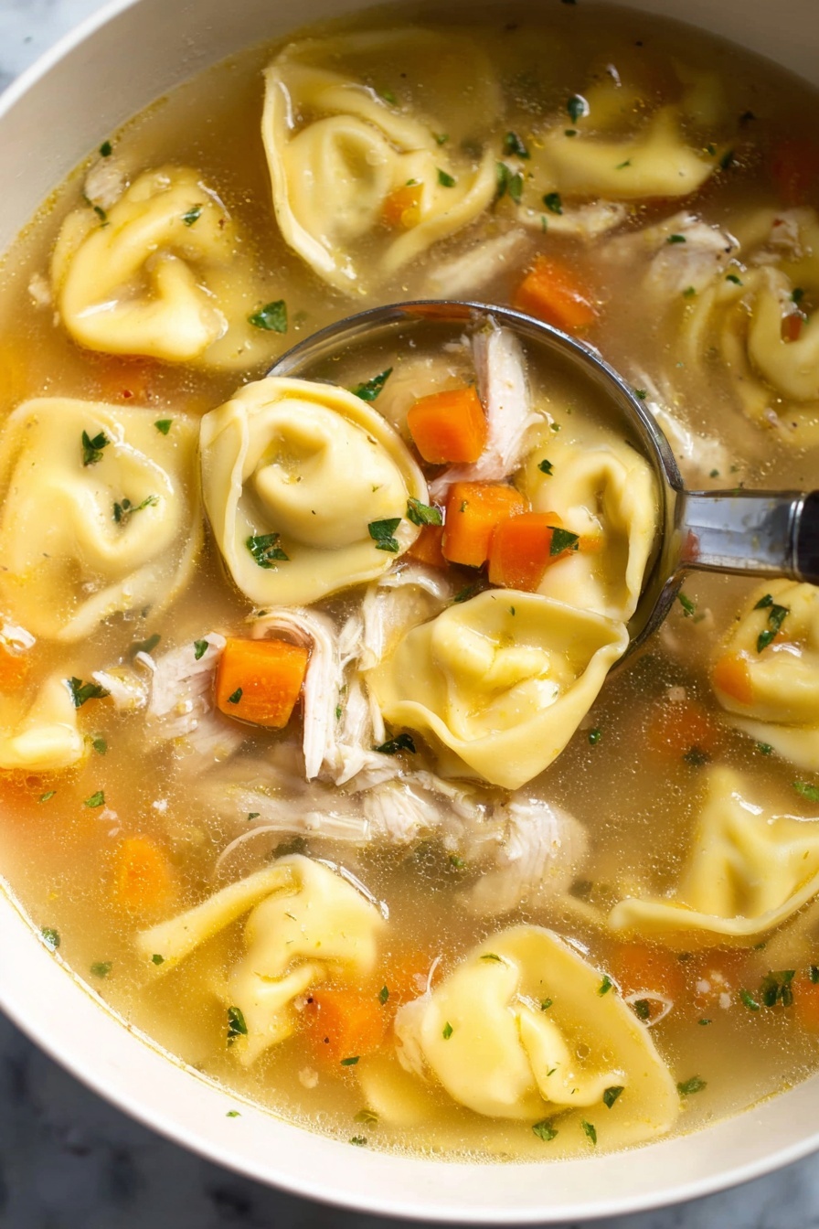 This image shows a close-up view of a bowl of clear broth soup filled with three main layers: the large, yellow tortellini pasta with a smooth, slightly wrinkled texture placed throughout; small, bright orange carrot cubes scattered evenly; and pieces of shredded white meat chicken mixed in. Small bits of green herbs float on top, adding a pop of color. A silver spoon with a long handle is scooping up the soup from the bowl, which is white, set on a white marbled surface. The broth has a light golden color and looks warm and inviting. Photo taken with an iphone --ar 2:3 --v 7 - Chicken Tortellini Soup, easy chicken tortellini soup, cheesy tortellini soup, comforting chicken soup recipe, quick weeknight soup