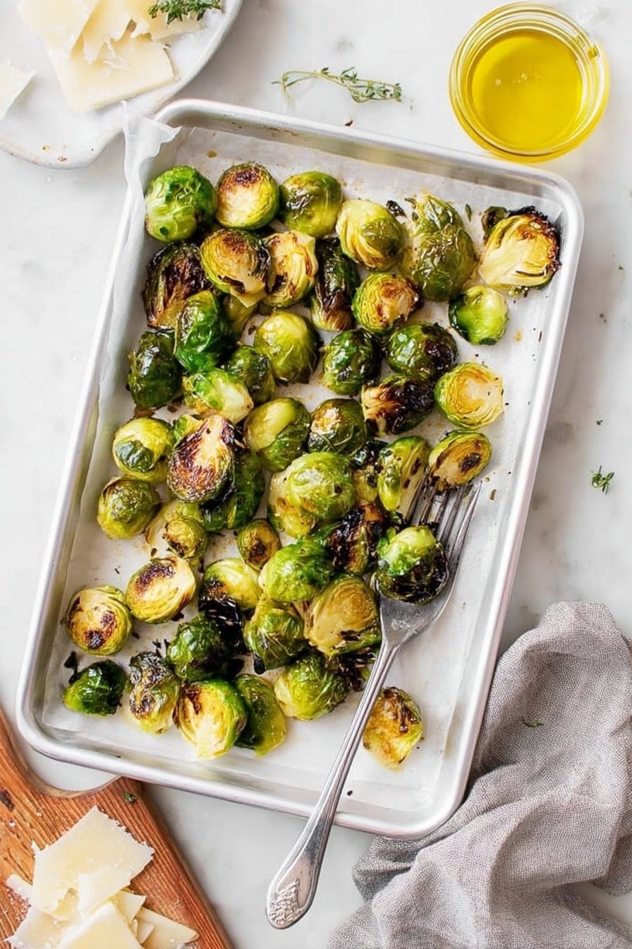 A white baking tray lined with white parchment paper holds a single layer of roasted Brussels sprouts, some halved and some whole, showing a mix of bright green and dark brown charred spots, giving a crispy texture. The tray is placed on a white marbled surface with a silver fork resting in the bottom right corner inside the tray. To the right, there is a small glass jar filled with yellow oil, and at the bottom left corner, part of a wooden board with thinly shaved white cheese is visible, along with a grey cloth nearby. The overall scene is bright and fresh, highlighting the crispy roasted Brussels sprouts. photo taken with an iphone --ar 2:3 --v 7 - Roasted Brussels Sprouts with Lemon, Parmesan, and Thyme, Brussels Sprouts Side Dish, Easy Roasted Brussels Sprouts, Lemon Parmesan Brussels Sprouts, Healthy Brussels Sprouts Recipe