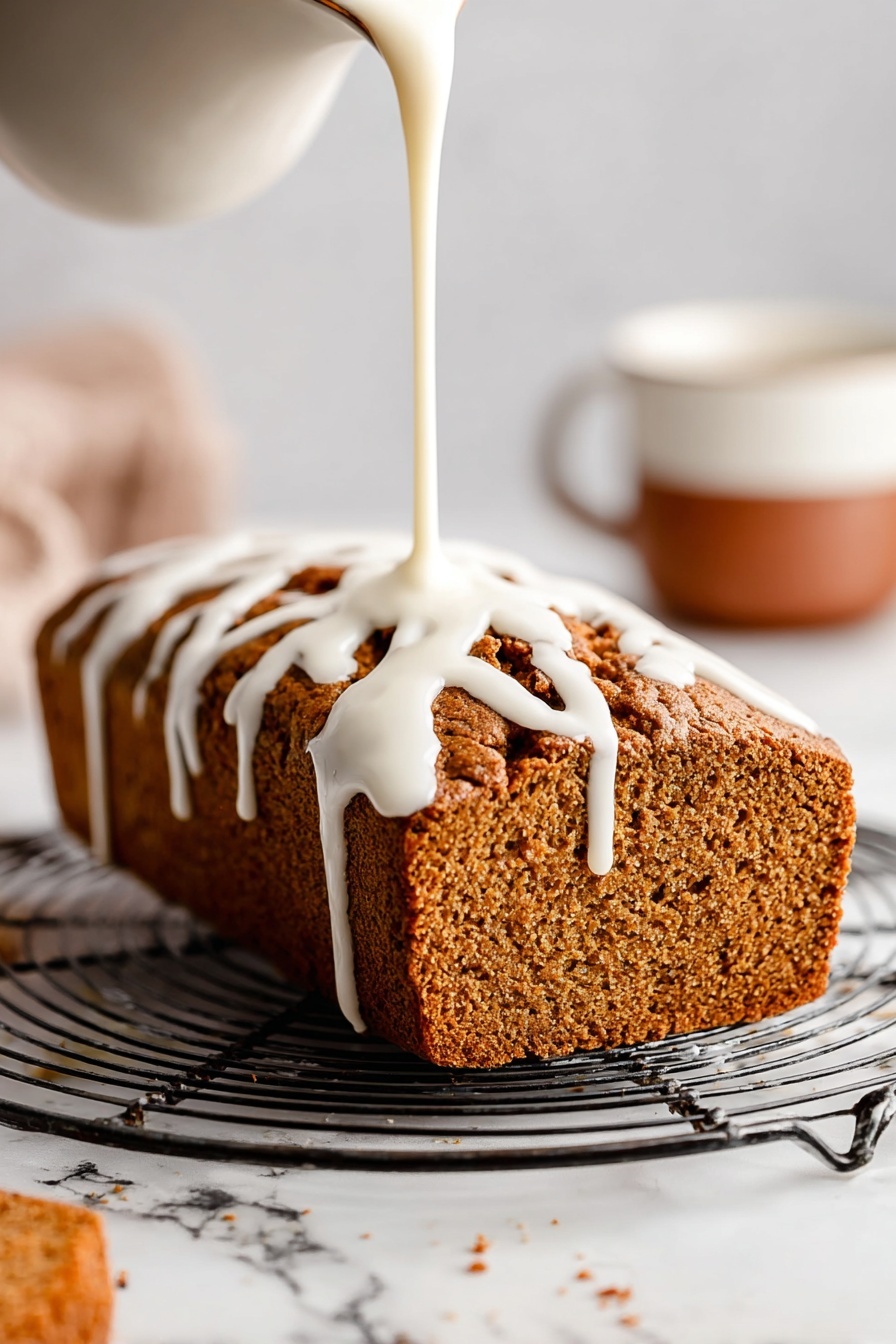 A loaf of brown textured bread sits on a round metal cooling rack with open grids. White icing is being poured over the top from above, flowing down the sides in thick, smooth ribbons. The loaf has a rough crust with deep cracks and a soft, porous interior visible on the side. In the background to the right, there is a blurred white cup with a brown lower half, all on a white marbled surface. Photo taken with an iphone --ar 2:3 --v 7 - Healthy Pumpkin Bread with Maple Glaze, healthy pumpkin bread, fall pumpkin bread recipe, nutritious pumpkin loaf, cozy spice pumpkin bread