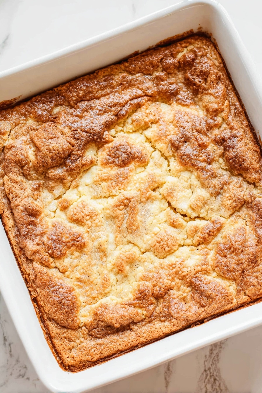 A square white baking dish holds a baked dessert with one thick layer that has a rough, cracked, golden-brown surface. The top looks crunchy and uneven with some lighter and darker brown spots, showing a slightly bubbled texture. The edges are more browned and crisp, while the center parts have a mix of smooth and bumpy patches. The dish is placed on a white marbled surface. Photo taken with an iphone --ar 2:3 --v 7 - Apple Dump Cake, easy apple dessert, quick apple cake, cinnamon apple crumble, simple fruit cobbler