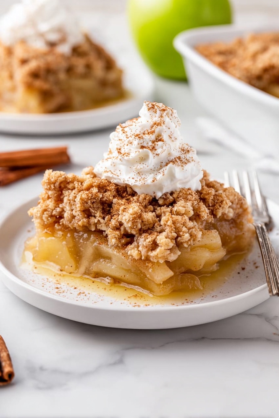 A slice of apple crumble sits on a white plate with three layers visible: a bottom layer of soft, cooked apple slices in a light golden sauce, a thick middle layer of crumbly, light brown streusel topping with a rough texture, and a dollop of white whipped cream sprinkled lightly with cinnamon on top. In the background, there is a second plate with more apple crumble and a silver fork. The scene is set on a white marbled surface with a halved green apple and a cinnamon stick placed nearby. Photo taken with an iphone --ar 2:3 --v 7 - Apple Dump Cake, easy apple dessert, quick apple cake, cinnamon apple crumble, simple fruit cobbler