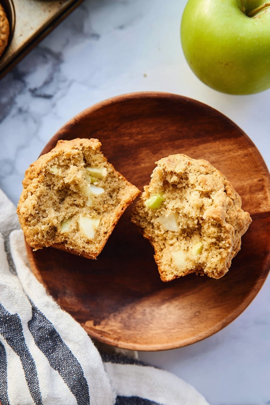A round wooden plate on a white marbled surface holds a split muffin showing its soft, light brown crumb mixed with small white and slightly green apple pieces inside. To the right, a green apple sits partially in frame, and a striped white and dark blue cloth is partly visible beneath the plate and muffin tray corner. The muffin has a slightly golden crust with a rough texture. photo taken with an iphone --ar 2:3 --v 7 - Healthy Apple Muffins, healthy apple muffins, nutritious apple muffin recipe, quick healthy muffins, apple cinnamon muffins