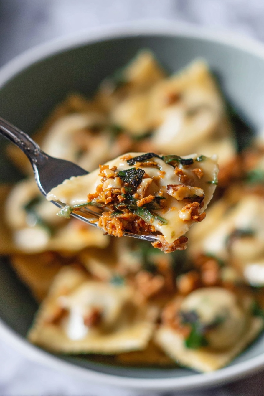 A close-up view shows a silver fork holding a piece of ravioli filled with a rich orange-brown meat mixture, topped with small dark green herb pieces and bits of light brown walnuts. In the blurred background, there is a white bowl filled with more ravioli, showing a mix of creamy beige pasta, white sauce, greens, and scattered walnut pieces, all set against a white marbled surface. The scene captures the soft texture of the ravioli and the fresh, rustic topping with a natural light that highlights the details. photo taken with an iphone --ar 2:3 --v 7 - Pumpkin Ravioli with Apple Sage Butter, autumn pasta recipes, homemade pumpkin ravioli, fall comfort food, sage butter pasta