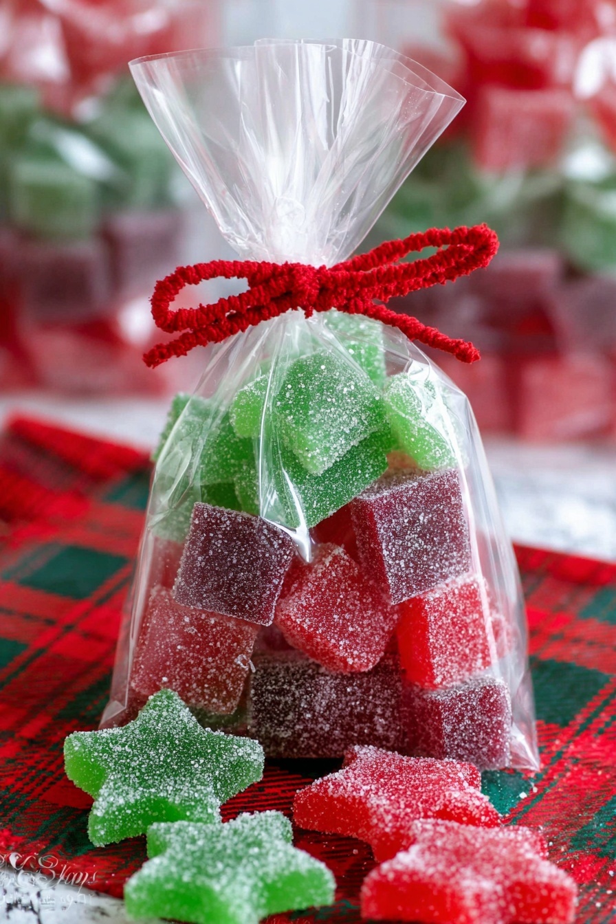 A clear plastic bag filled with stacked square sugar-coated gummies in red, green, and purple colors, arranged in alternating layers. There are also red and green star-shaped sugar-coated gummies at the bottom of the bag and scattered outside on a red and green plaid cloth. The bag is tied at the top with a red twist ribbon and decorated with a red felt scalloped band. The background shows a white marbled texture with blurred additional bags of similar candy. Photo taken with an iphone --ar 2:3 --v 7 - Homemade Gelatin Gumdrops, easy homemade candy, chewy fruit gums, DIY gelatin candies, natural gummy treats