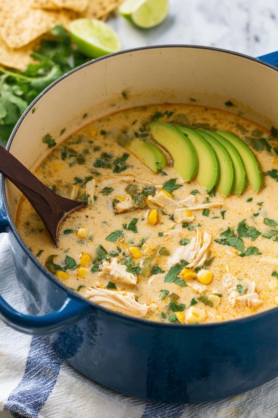 A large blue pot filled with creamy soup that has a light orange color with green cilantro leaves scattered on top. Inside the soup, there are chunks of white chicken, pieces of yellow corn, and bits of green vegetables. On one side, thin slices of light green avocado are placed in a neat row, partially submerged in the soup. A dark wooden spoon is stirring the soup from the left side. The pot sits on a white cloth with blue stripes on a white marbled surface. In the background, there are some light beige tortilla chips and lime wedges. Photo taken with an iphone --ar 2:3 --v 7 - White Chicken Chili, easy white chicken chili, creamy chicken chili, quick chicken chili recipe, comforting white chili