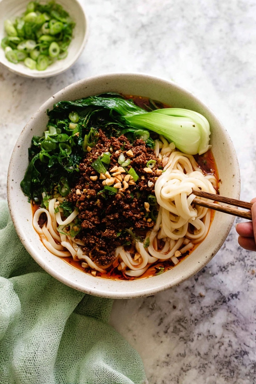 A white bowl filled with three main layers: at the bottom, thick white noodles mixed with a red oily sauce that gives a shiny texture; on top, a layer of dark brown minced meat scattered evenly; around the edges, bright green leafy vegetables adding fresh color; garnished with sliced light green scallions and small beige crushed nuts. A pair of dark brown chopsticks hold a small twisted bunch of noodles dipped in the sauce at the right side of the bowl. Some scallions and nut bits are scattered on the dark surface around the bowl. photo taken with an iphone --ar 2:3 --v 7 - Spicy Dan Dan Noodles with Pork, Sichuan Dan Dan Noodles, Easy Spicy Pork Noodles, Authentic Dan Dan Noodles Recipe, Savory Pork Noodle Bowl