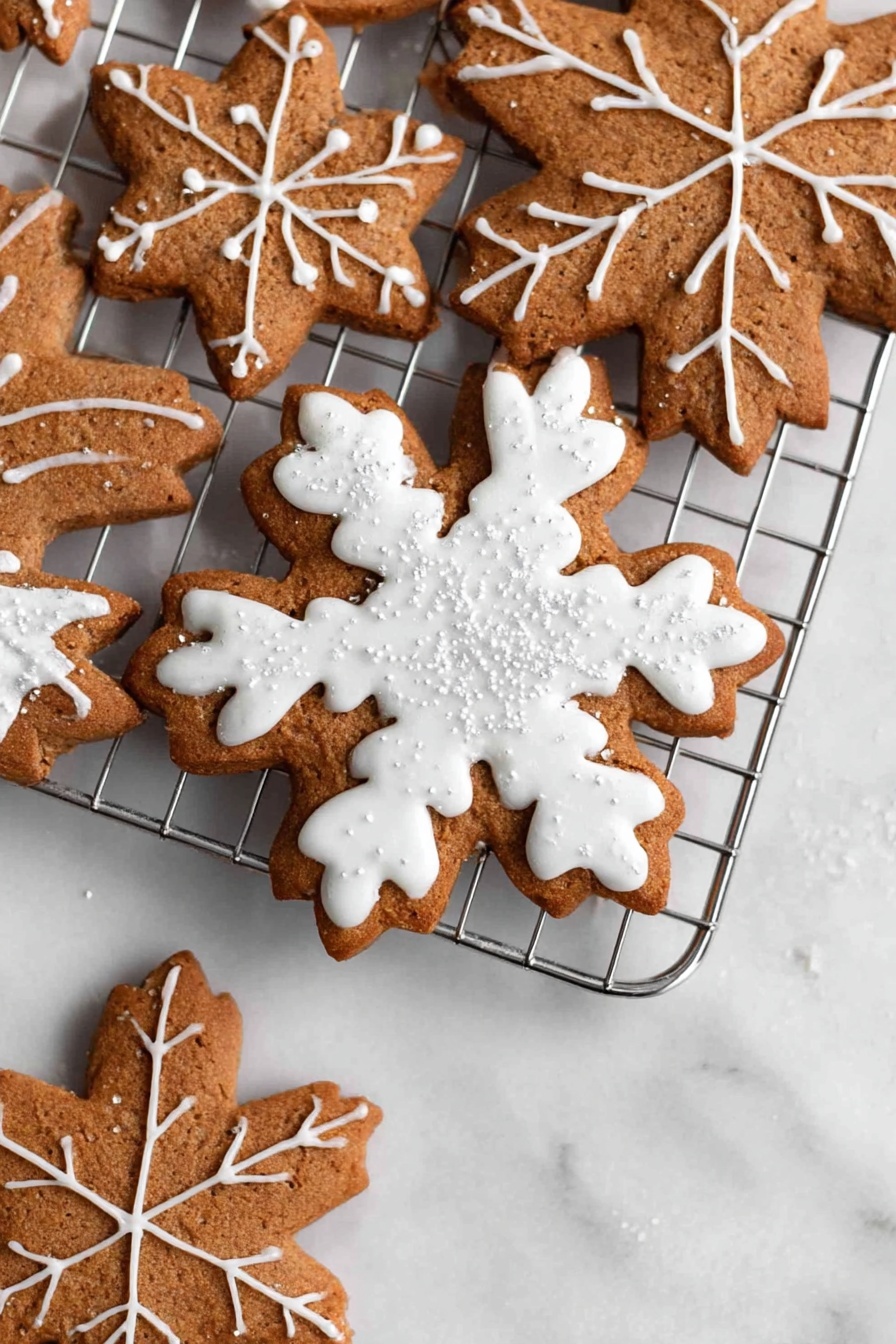 The image shows three snowflake-shaped cookies stacked on a silver cooling rack placed on a white marbled surface. The cookies are brown with detailed white icing decoration. The top cookie has thick white icing covering the shape with a leaf vein pattern on each arm and small shiny sugar crystals scattered on top. The two cookies underneath have thinner white icing lines creating a delicate snowflake design with crisscross patterns. The cookies’ texture looks soft yet firm with visible edges. Photo taken with an iphone --ar 2:3 --v 7 - Vegan Gingerbread Cookies with Icing, plant-based holiday cookies, vegan Christmas cookies, gluten-free gingerbread cookies, vegan festive baking
