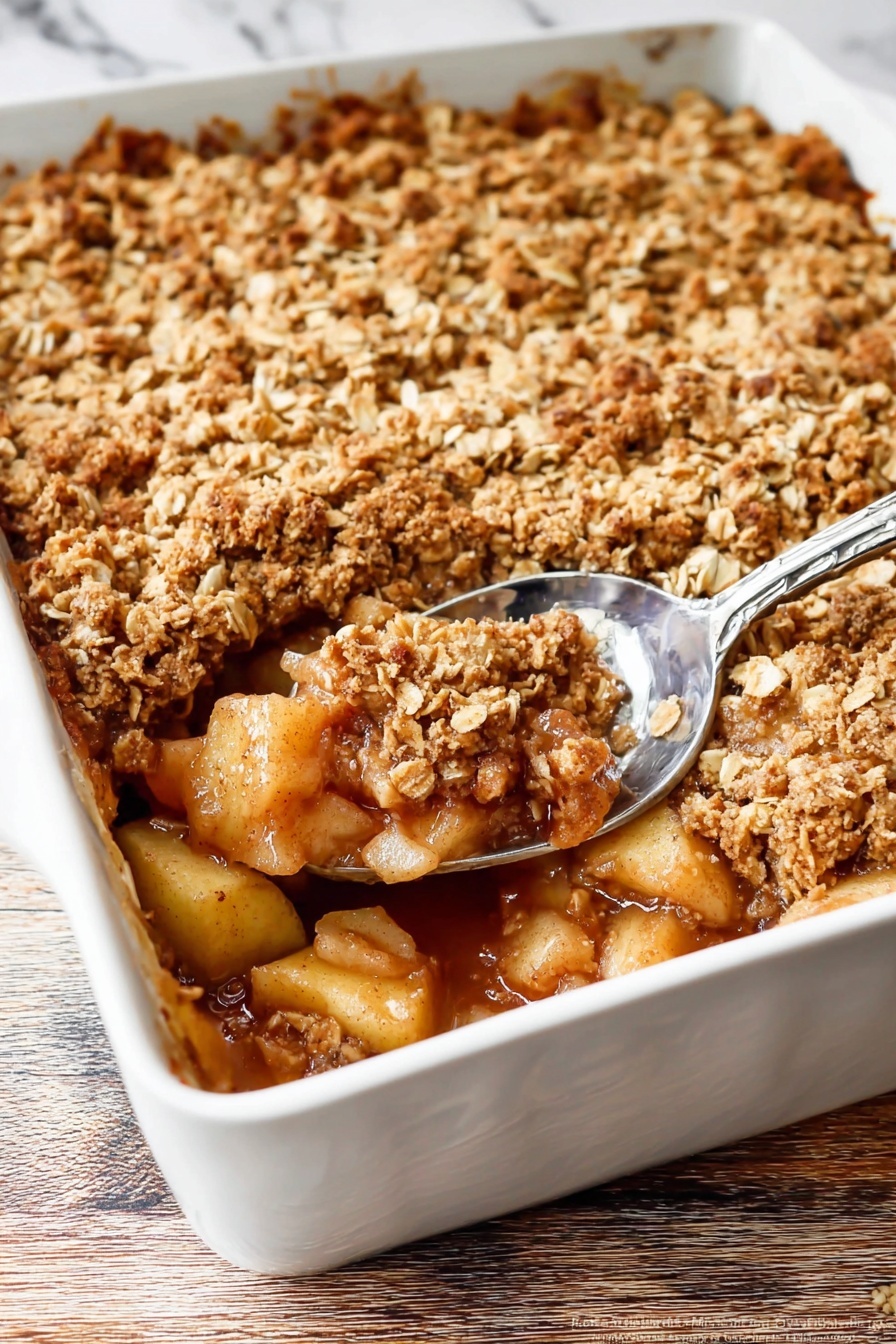 The image shows a white baking dish filled with a baked apple dessert. The bottom layer consists of soft, cooked apple chunks in a golden-brown syrup. Above this is a thick layer of crumbly oat topping, golden and slightly toasted with visible oats and brown sugar. A silver spoon scoops out some of the dessert from the bottom right corner, revealing the juicy apple filling beneath the crunchy topping. The white dish is set on a wooden surface with a white marbled texture in the background. Photo taken with an iphone --ar 2:3 --v 7 - Easy Classic Apple Crisp, apple crisp dessert, easy apple dessert, comforting apple crisp, simple apple crumble