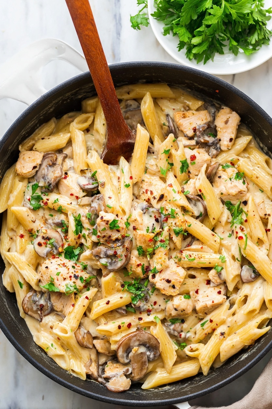 A black pan filled with creamy pasta made of three main layers: the base layer of pale yellow penne pasta, the middle layer of light brown cooked mushroom slices, and the top layer of small light beige pieces of chicken mixed in a creamy sauce. Green parsley leaves are scattered on top, adding bright green spots, with tiny black and red pepper flakes sprinkled over all. A wooden spoon rests inside the pan, partially covered by the pasta. The pan is placed on a white marbled surface, with a small white plate of fresh parsley leaves in the top corner. photo taken with an iphone --ar 2:3 --v 7 - Creamy Chicken Marsala Pasta, Chicken Marsala pasta recipe, easy chicken Marsala, creamy pasta with Marsala wine, comforting chicken pasta dish