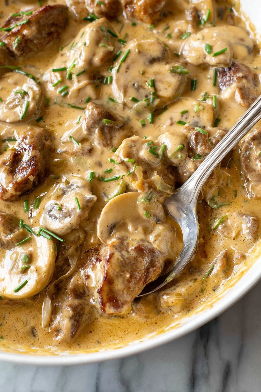 This image shows a close-up of a creamy dish with several layers visible. The base layer is a thick, light brown sauce with a smooth, slightly shiny texture, pooling around the other ingredients. Mixed in the sauce are slices of light beige mushrooms with soft, rounded edges and a slightly wet look. Also present are pieces of browned meat, which have a slightly rough texture and are coated in the same sauce. Small green chive pieces are scattered on top, adding little spots of bright green color throughout. A silver spoon is partially dipped into the dish, resting on the edge of a white bowl. The background is a white marbled surface. Photo taken with an iphone --ar 2:3 --v 7 - Beef Stroganoff, Beef Stroganoff with Mushrooms, Creamy Beef Stroganoff, Classic Beef Stroganoff, Easy Beef Stroganoff