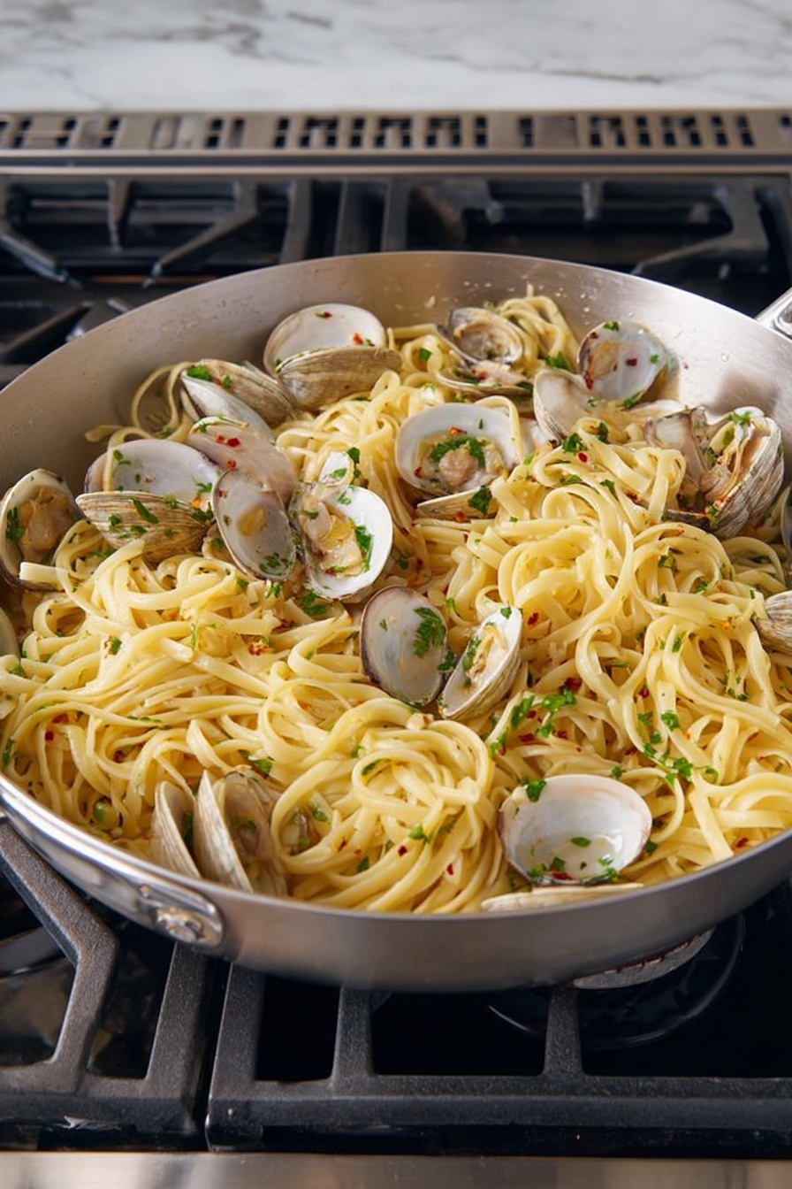 A large silver pan filled with cooked linguine pasta mixed with open clams distributed evenly throughout. The pasta is light yellow, soft and slightly shiny from sauce, with chopped green herbs sprinkled on top for color and freshness. Small bits of garlic and red chili flakes are visible mixed into the pasta, adding texture and flavor. The pan sits on a black stove with metal grates, surrounded by a white marbled surface. photo taken with an iphone --ar 2:3 --v 7 - Linguine with Clams and Lemon, seafood pasta with lemon, quick seafood dinner, Italian clam pasta, easy lemon linguine