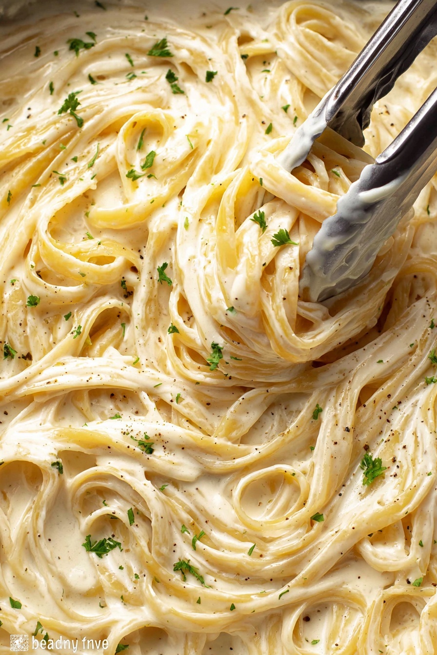 A close-up view of creamy fettuccine pasta in a thick white sauce filling the whole frame, with the pasta strands twisted and intertwined. The sauce is smooth and glossy with a light beige color and flecks of black pepper and small green parsley pieces sprinkled on top. Silver tongs are grabbing a bunch of pasta near the center, showing the rich texture of the sauce coating the noodles. The background is a white marbled texture. photo taken with an iphone --ar 2:3 --v 7 - Creamy Chicken Alfredo Pasta, easy chicken Alfredo, homemade Alfredo sauce, creamy pasta recipes, family-friendly pasta dishes