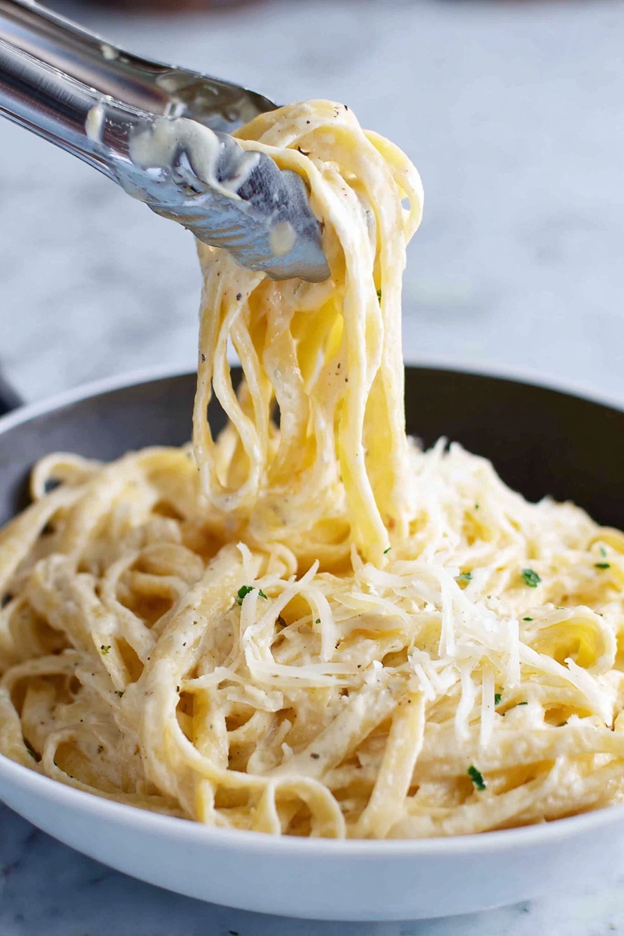 A close-up image shows thick noodles coated with a creamy white sauce, twirled and lifted by silver tongs above a white bowl filled with the same noodles. The noodles have a slightly glossy texture from the sauce and small green herb bits sprinkled throughout. Light shreds of pale cheese are scattered on top, adding a soft, fine texture contrast against the smooth noodles. The background is a white marbled surface. photo taken with an iphone --ar 2:3 --v 7 - Fettuccine Alfredo, Alfredo with Parmesan, creamy pasta, homemade Alfredo sauce, classic pasta dishes