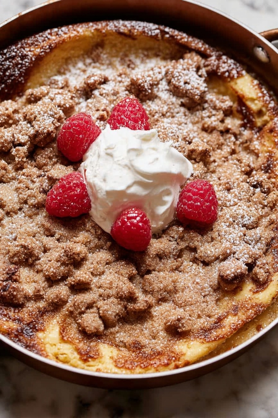 A round dessert with two main layers is shown in a pan. The bottom layer is a golden-brown, slightly risen cake with some darker browned spots. On top is a crumbly, light brown streusel layer that covers most of the surface unevenly. There is a dollop of white cream in the center with four bright red raspberries placed around and on it. A light dusting of powdered sugar is scattered over parts of the streusel and cake edges. The pan sits on a white marbled surface. photo taken with an iphone --ar 2:3 --v 7 - Brioche French Toast Casserole, breakfast casserole with brioche, decadent French toast bake, easy brunch casserole, cinnamon streusel French toast