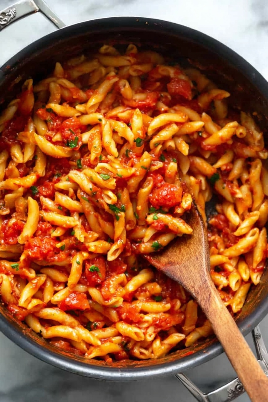 A black pot filled with a single layer of short, twisted pasta all covered evenly in a bright red tomato sauce with small bits of tomatoes and green herbs mixed in. A woman’s wooden spoon is stuck deep in the middle, lifting some pasta, showing the sauce clinging to the noodles. The pot is on a white marbled surface, the pasta looks soft and shiny with sauce. Photo taken with an iphone --ar 2:3 --v 7 - Spicy Penne Arrabbiata, Penne Arrabbiata sauce, Italian pasta recipes, easy spicy pasta, quick dinner ideas