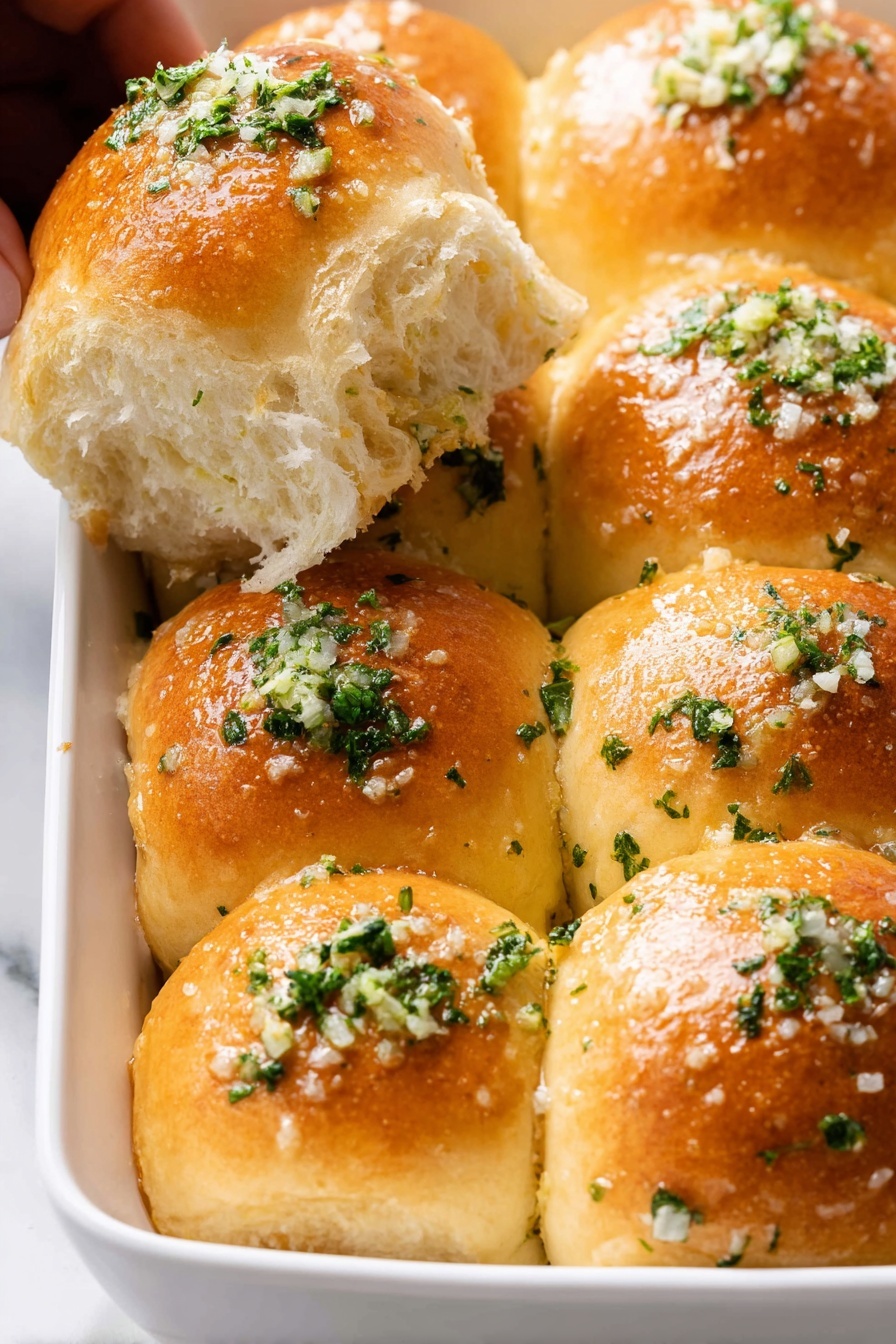 The image shows a white dish filled with seven shiny, golden-brown dinner rolls bunched together in two rows. Each soft roll is topped with small bits of green parsley and finely chopped garlic, sprinkled evenly with coarse salt. One roll is slightly pulled away, revealing a fluffy, light inside with a soft texture. There is a woman's hand partially holding this separated roll on the top left of the frame. The background and surface are a white marbled texture. photo taken with an iphone --ar 2:3 --v 7 - Garlic Bread Rolls for Thanksgiving, Thanksgiving bread side dish, cheesy garlic dinner rolls, soft buttery garlic rolls, holiday bread recipes