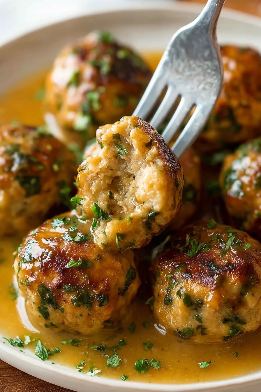 The image shows several round meatballs with a golden-brown crust and green herb bits scattered through them, resting in a light brown gravy. A silver fork holds one meatball with a bite taken out, showing a soft and moist inside with visible herbs. The meatballs are arranged closely on a white plate with some chopped green herbs sprinkled on top and the gravy pooling around them. The background is a white marbled texture photo taken with an iphone --ar 2:3 --v 7 - Stuffing Balls, holiday stuffing ideas, savory stuffing bites, Thanksgiving appetizers, baked stuffing balls