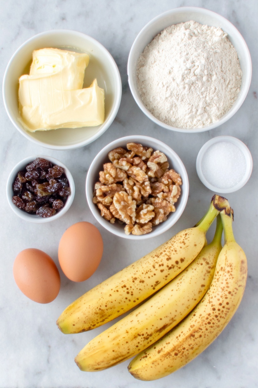 Flat lay of three very ripe bananas with brown speckles, a small white ceramic bowl filled with softened unsalted butter, another white bowl containing granulated sugar, two whole uncracked brown eggs, a white bowl with all-purpose flour, a small white bowl holding walnuts roughly chopped, a small white bowl filled with dark raisins, a small white bowl with pale beige baking soda powder, a tiny white bowl with coarse salt, and a small white bowl with golden vanilla extract, all arranged with perfect symmetry on a clean white marble surface, soft natural light, photo taken with an iPhone, professional food photography style, fresh ingredients, white ceramic bowls, no bottles, no duplicates, no utensils, no packaging --ar 2:3 --v 7 --p m7354615311229779997 - Moist Banana Bread with Walnuts and Raisins, banana bread recipe, easy banana bread, healthy banana bread, best banana loaf