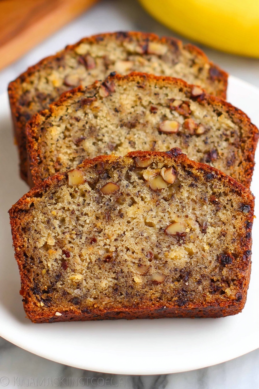 Three slices of banana nut bread are stacked slightly overlapping on a white plate, showing a dense texture filled with dark brown nuts and small chunks of banana inside the golden brown cake. The crust is a darker shade of brown and slightly crisp, while the inside looks moist with a mix of light and dark brown colors from the bananas and nuts. The plate is on a white marbled surface with a blurred yellow banana visible in the background. Photo taken with an iphone --ar 2:3 --v 7 - Moist Banana Bread with Walnuts and Raisins, banana bread recipe, easy banana bread, healthy banana bread, best banana loaf