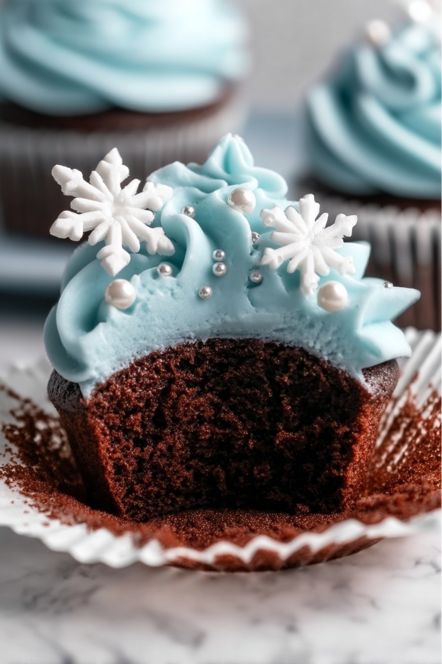 A close-up view of a chocolate cupcake with a white cupcake liner partially peeled back, showing the soft, dark brown cake inside. The top layer is covered with smooth, light blue frosting piped in swirls, decorated with small silver pearls and two white snowflake-shaped sprinkles placed on the side. The cupcake is placed on a white marbled surface. Photo taken with an iphone --ar 2:3 --v 7 - Winter Blue Chocolate Cupcakes with Snowflake Frosting, festive winter cupcakes, holiday chocolate cupcakes, snowflake frosting recipe, blue buttercream cupcakes