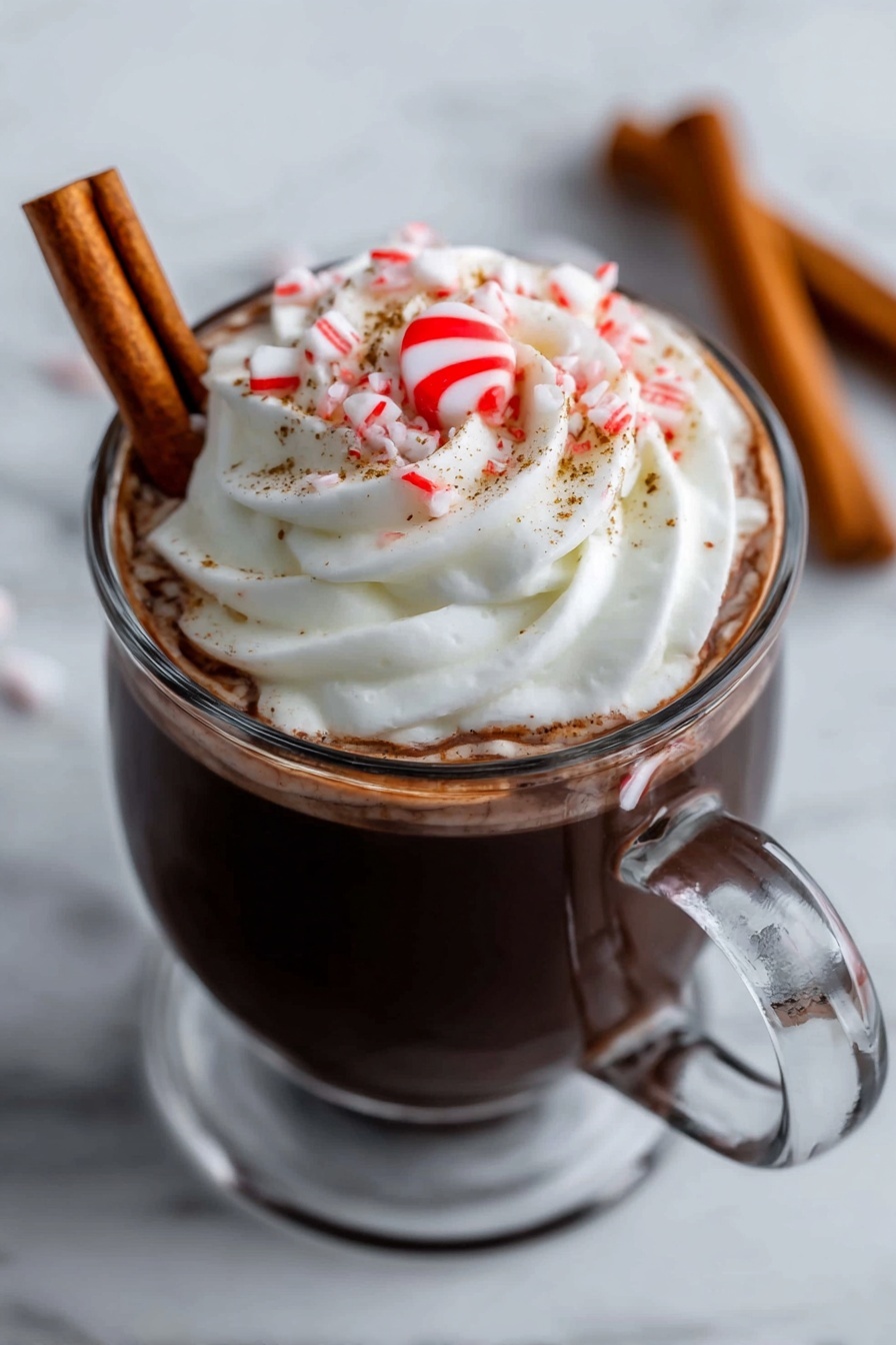 A clear glass mug filled with dark hot chocolate topped with a large swirl of white whipped cream. On top of the cream is a small red and white peppermint candy in the center, surrounded by some small pieces of crushed candy cane and fine brown sprinkles. A brown cinnamon stick is resting against the side inside the mug. The mug is placed on a white marbled surface. In the background, two more cinnamon sticks lie on the surface out of focus. A woman's hand is holding the handle of the mug. photo taken with an iphone --ar 2:3 --v 7 - Festive Peppermint Hot Chocolate, holiday hot chocolate recipe, peppermint hot cocoa, winter hot chocolate ideas, creamy peppermint hot chocolate