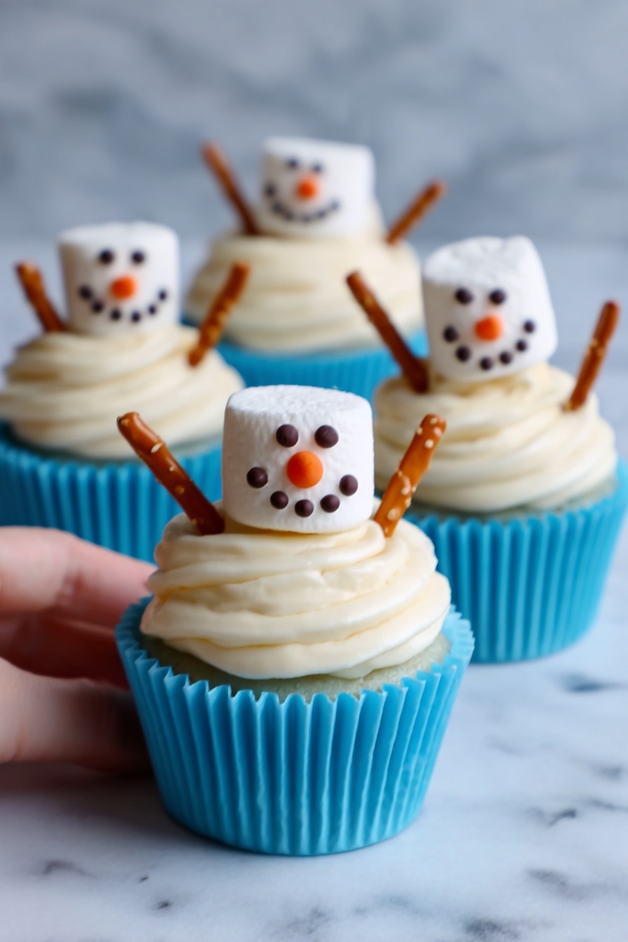 The image shows three cupcakes in white cupcake liners with bright blue bases, each topped with thick, creamy white frosting swirled in layers. On top of each cupcake sits a large marshmallow decorated like a snowman face with small dark chocolate or icing dots for eyes and mouth, and an orange triangular nose. Two pretzel sticks are inserted on opposite sides of the frosting, looking like the snowman's arms. The cupcakes are placed on a white marbled surface, with one woman's hand gently holding the front cupcake. The scene has bright, soft lighting. photo taken with an iphone --ar 2:3 --v 7 - Snowman Cupcake Muffins, festive holiday cupcakes, winter dessert ideas, chocolate muffin recipes, kids holiday treats