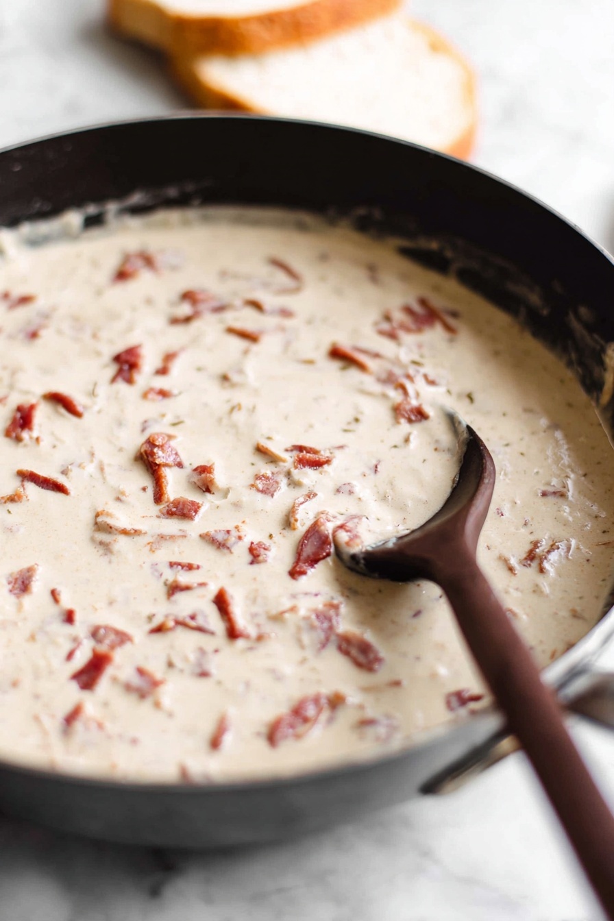 A white plate with ridged edges holds two slices of golden toasted bread stacked slightly off-center, covered generously with a thick, creamy white sauce that has visible small chunks of reddish-brown bacon and tiny bits of green herbs sprinkled on top. The sauce drips slightly over the edges of the toast, and a few small crumbs and herbs are scattered around the plate. The plate rests on a white marbled surface. photo taken with an iphone --ar 2:3 --v 7 - Chipped Beef on Toast, classic comfort food, easy breakfast recipes, hearty quick meals, savory breakfast ideas