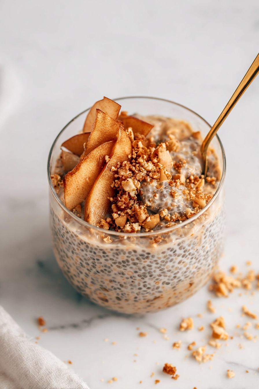 A clear glass filled with three main layers is shown on a white marbled surface. The bottom layer is a thick, creamy mixture with visible small black chia seeds evenly distributed, giving it a speckled texture. On top of that, there is a layer of thin, light brown apple slices with a soft texture, slightly curled and overlapping in a loose arrangement. The top layer is sprinkled with small, coarse granules of golden brown crumbs and chopped nuts, adding a crunchy texture and contrasting color. A gold spoon is inserted on the right side of the glass, partly immersed in the layers. Some crumbs are scattered around on the white marbled surface near the glass, showing slight messiness. photo taken with an iphone --ar 2:3 --v 7 - Apple Cinnamon Chia Pudding with Crispy Crumble, healthy breakfast chia pudding, apple cinnamon overnight oats, nourishing chia pudding recipes, easy fruit crumble desserts