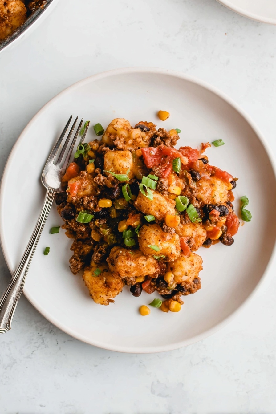 A white plate sits on a white marbled surface, holding a layered dish. The bottom layer includes browned, small potato puffs that are crispy. Mixed in are bits of yellow corn and black beans, creating specks of yellow and black. There are pieces of green onions scattered on top, adding a fresh green color. Red tomato pieces and small bits of a brown sauce or meat mixture cover the dish, giving it a rich texture. A silver fork rests on the left side of the plate partly under the food. Photo taken with an iphone --ar 2:3 --v 7 - Cowboy Tater Tot Casserole, Tater Tot Casserole, cheesy meaty casserole, easy comfort food, hearty weeknight dinner