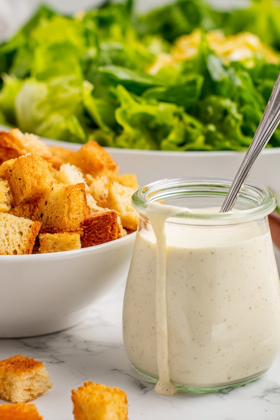 The image shows a close-up of a small clear glass jar filled with thick, creamy white dressing with tiny brown specks, some of which is dripping down the side. A metal spoon sticks out from the jar. To the left, there is a white bowl filled with golden brown croutons, varying in shades and textures, some smooth and some rough. In the background, a large white bowl holds bright green leafy lettuce, showing fresh, wavy textures. The surface beneath everything is a white marbled texture. photo taken with an iphone --ar 2:3 --v 7 - Creamy Lemon Salad Dressing, lemon salad dressing recipe, tangy salad dressing, quick healthy salad dressing, versatile lemon dressing
